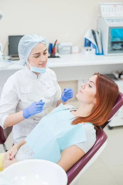 Giovane bella dentista femminile aiuta paziente con mal di denti. Una donna dai capelli rossi siede su una sedia dal medico e si lamenta del dolore della carie. Servizi dentistici. Ragazza infelice con pulpite — Foto Stock