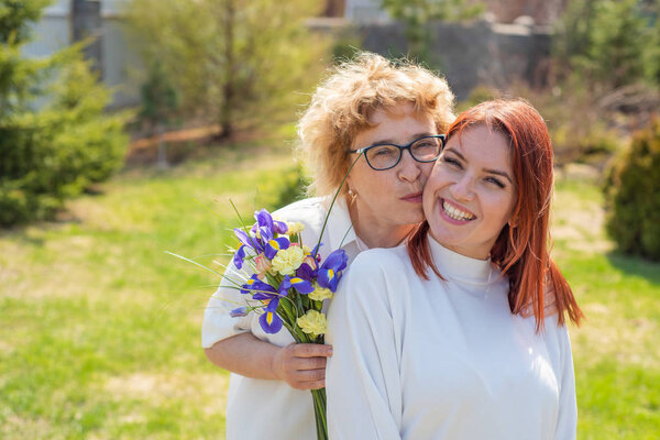Взрослая дочь дарит цветы своей матери на улице, во дворе дома. Проводим время вместе, празднуем дома по выходным. День Матери. Теплые отношения между поколениями
