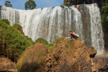 Gözlüklü genç ve kızıl saçlı genç bir kadın, şelalenin yakınındaki bir kayalıkta titibhasana pozuyla ellerinde duruyor. Kız bir ateş böceği poz doğada yoga uygulamaları. Vahşi doğada pilates.