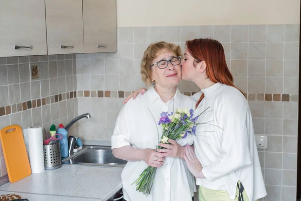 Η κόρη δίνει λουλούδια στην ενήλικη μητέρα της και την φιλάει στην κουζίνα. Περνάμε χρόνο μαζί, γιορτάζοντας στο σπίτι τα Σαββατοκύριακα. Ημέρα της μητέρας. Δάκρυα ευτυχίας. — Φωτογραφία Αρχείου