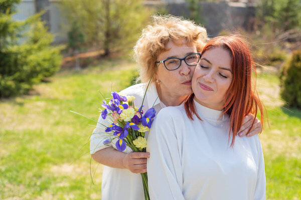 Взрослая дочь дарит цветы своей матери на улице, во дворе дома. Проводим время вместе, празднуем дома по выходным. День Матери. Теплые отношения между поколениями
