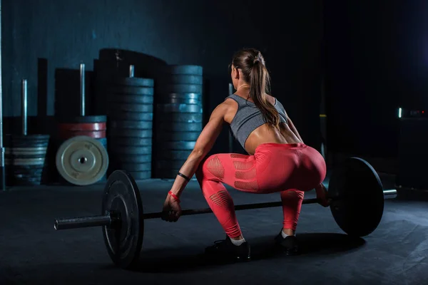 赤いレギンスの若い、ヨーロッパの、筋肉の女の子は、クロスフィットのためのジムでバーベルで運動をしています。暗い背景 — ストック写真