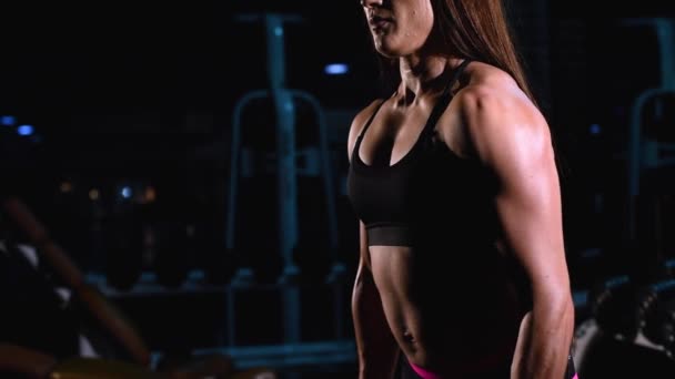 Forte, musclée, la fille en short noir et un haut, entraîne un biceps d'une main, lèvera des haltères de sport. Le gymnase sombre . — Video