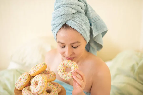 A rapariga de toalha toma o pequeno-almoço na cama. Uma jovem mulher atraente come donuts na cama. A menina com uma toalha na cabeça come donuts alegremente. . — Fotografia de Stock