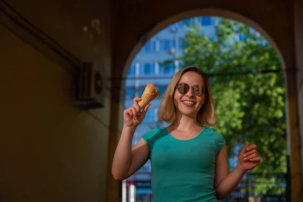 Uma mulher feliz em um vestido turquesa fica em uma passagem arqueada e come um cone de waffle em um dia quente de verão. Linda loira em óculos de sol gosta de sorvete enquanto caminha. Sobremesa de verão. — Fotografia de Stock