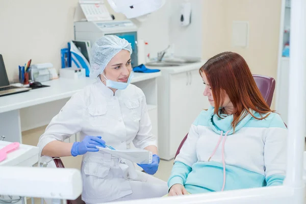 Giovane bella dentista che parla con una paziente in studio. Una donna dai capelli rossi siede su una sedia con un ortodontista e ascolta gli appuntamenti. Servizi dentistici — Foto Stock