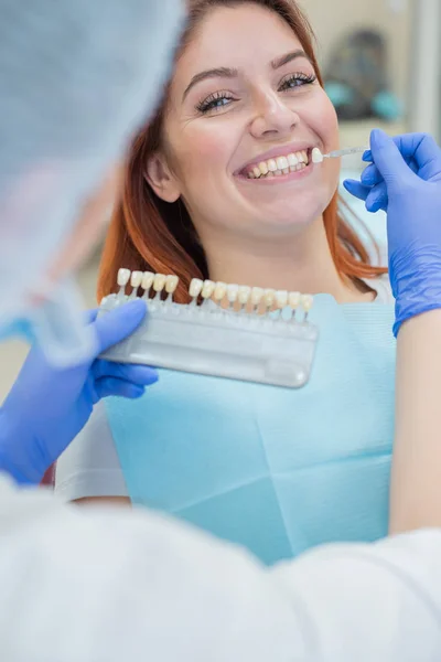 Controllare e selezionare il colore dei denti sulla sedia del dentista. Il dentista fa il processo di trattamento di una bella giovane ragazza dai capelli rossi. Selezione di un impianto dentale . — Foto Stock
