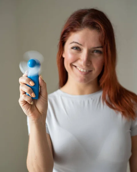 Ritratto di una donna dai capelli rossi sorridente con un ventilatore tascabile in mano. Faccia piena di una ragazza rinfrescante nel calore di un colpo di vento da un dispositivo elettrico wireless. Flusso d'aria fresca dal condizionatore — Foto Stock