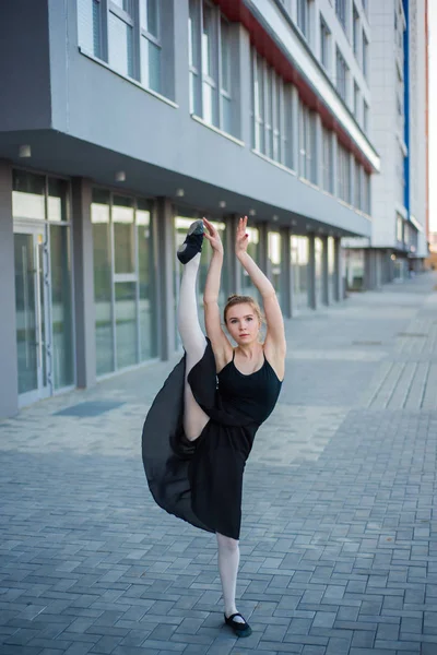 Ballerina in tutu posing against a residential building. Beautiful young woman in a black dress and pointe shoes jumping demonstrates stretching. Ballerina is standing in the splits. Outside. — Stock Photo, Image