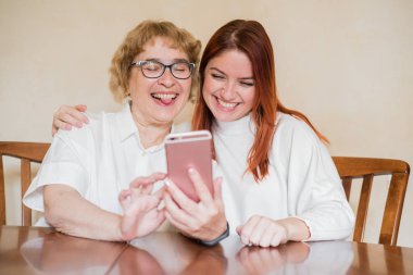 Anne ve kızı masada evde otururken akıllı telefonun ön kamera fotoğraflandı. Neşeli yaşlı anne ve kızı komik selfie'ler yapıyor ve dillerini gösteriyor.