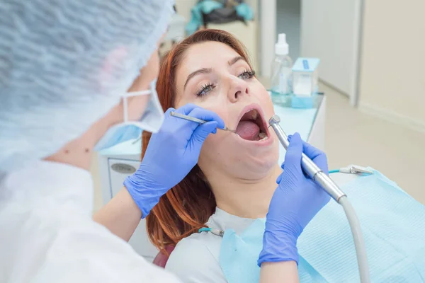 Una atractiva doctora sonriente en uniforme blanco examina a una paciente de cabello rojo. Dentista produce trata los dientes con un taladro . — Foto de Stock