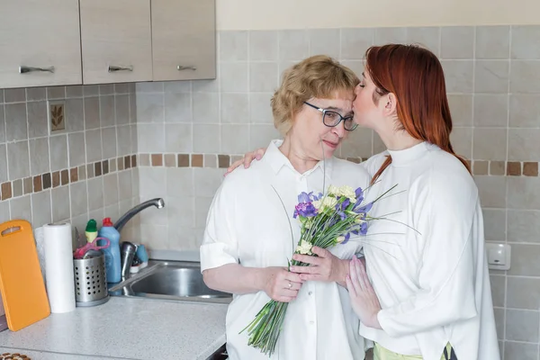 Η κόρη δίνει λουλούδια στην ενήλικη μητέρα της και την φιλάει στην κουζίνα. Περνάμε χρόνο μαζί, γιορτάζοντας στο σπίτι τα Σαββατοκύριακα. Ημέρα της μητέρας — Φωτογραφία Αρχείου