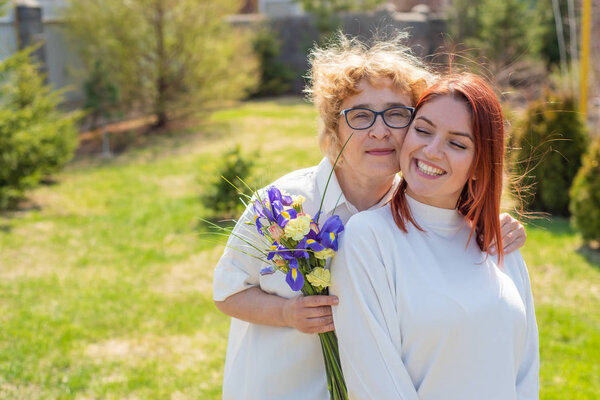 Взрослая дочь дарит цветы своей матери на улице, во дворе дома. Проводим время вместе, празднуем дома по выходным. День Матери. Теплые отношения между поколениями

