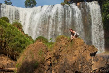 Gözlüklü genç ve kızıl saçlı genç bir kadın, şelalenin yakınındaki bir kayalıkta titibhasana pozuyla ellerinde duruyor. Kız bir ateş böceği poz doğada yoga uygulamaları. Vahşi doğada pilates.