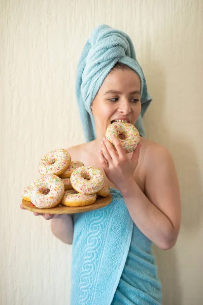 Flickan i en handduk har frukost i sängen. En ung attraktiv kvinna äter munkar i sängen. Flickan med en handduk på hennes huvud glatt äter munkar. — Stockfoto