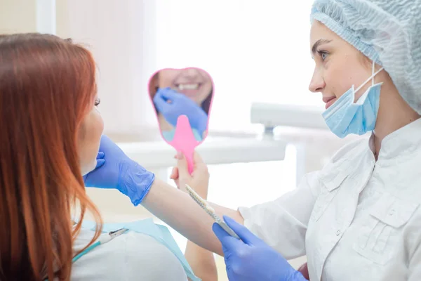 Controllare e selezionare il colore dei denti sulla sedia del dentista. Il dentista fa il processo di trattamento di una bella giovane ragazza dai capelli rossi. Selezione di un impianto dentale.Il riflesso nello specchio . — Foto Stock