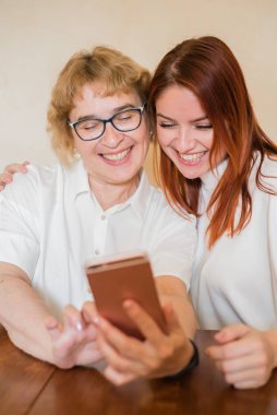 Hafif kıyafetler giymiş yaşlı bir anne ve kızı gülümsüyor ve selfie çekiyor. Anne ve kızı masada evde otururken akıllı telefonun ön kamera fotoğraflandı.