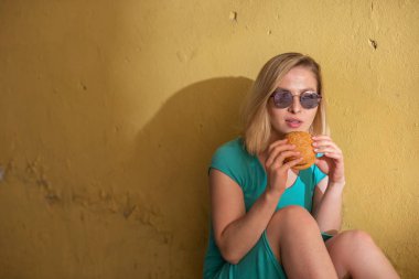 Turkuaz bir elbise içinde mutlu bir kadın kaldırımda oturur ve sıcak bir yaz gününde bir burger yiyor. Güneş gözlüğü güzel sarışın bir konut binanın sarı duvarında önemsiz yiyecek sahiptir.