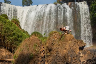 Gözlüklü genç ve kızıl saçlı bir kadın şelalenin yanındaki bir uçurumun üzerinde bokasane'de duruyor. Kız bir karga poz doğada yoga uygulamaları. Vahşi doğada pilates