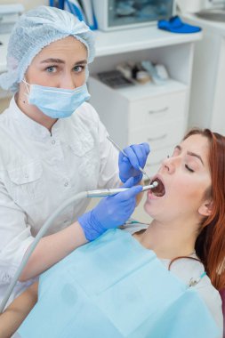 Beyaz üniformalı çekici, gülümseyen kadın doktor kızıl saçlı bir kadın hastayı muayene eder. Diş hekimi bir matkap ile diş davranır üretir.