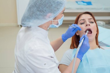 Beyaz üniformalı çekici, gülümseyen kadın doktor kızıl saçlı bir kadın hastayı muayene eder. Diş hekimi bir matkap ile diş davranır üretir.