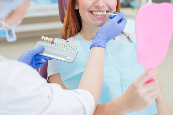 Controllare e selezionare il colore dei denti nella sedia dentista. Il dentista fa il processo di trattamento di una bella ragazza dai capelli rossi. Selezione di un impianto dentale. Il paziente si guarda allo specchio . — Foto Stock