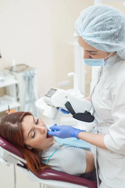 Atractiva doctora sonriente en uniforme blanco con herramientas en la mano. Joven dentista hermosa toma rayos X de mandíbula de paciente femenina. Mujer atractiva paciente en el dentista que toma examen de rayos X . — Foto de Stock