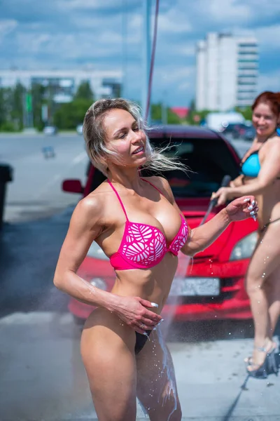 Chicas de lujo en bikinis seductores lavan un coche en un lavado de coches de la calle y el agua entre sí. Rubia y pelirroja con hermosas figuras lavan un coche rojo en un lavadero manual. . —  Fotos de Stock