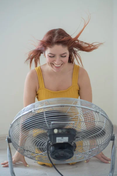 Portrait of a beautiful red-haired woman in a mustard jumpsuit enjoying the cooling breeze from a large electric fan. A smiling girl freshens up in the hot summer heat. Hair develops from the wind