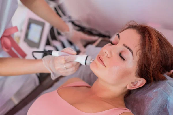 Giovane donna rossa ottiene un massaggio facciale elettrico nella lotta contro l'invecchiamento. La donna di 30 anni riceve il massaggio facciale ultrasonico elettrico contro rughe in un salone di bellezza. Cosmetologia hardware. — Foto Stock