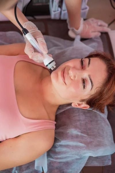 Joven pelirroja recibe un masaje facial eléctrico en la lucha contra el envejecimiento. Mujer de 30 años recibe masaje facial ultrasónico eléctrico contra arrugas en un salón de belleza. Cosmetología del hardware. — Foto de Stock