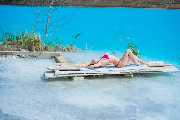 A beautiful red-haired woman in a seductive crimson bikini sits in front of an unusual turquoise lake. A miracle of nature, bright blue clear water. Ash dump, poisoned pond. — Stock Photo, Image