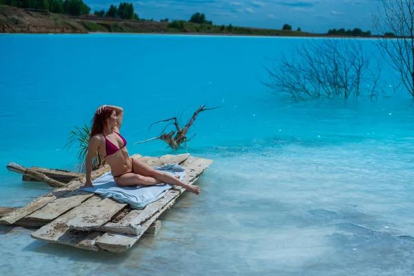 Une belle femme aux cheveux roux en bikini cramoisi séduisant se trouve en face d'un lac turquoise insolite. Un miracle de la nature, une eau claire bleu vif. Benne à cendres, étang empoisonné . — Photo