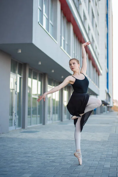 Ballerina in a tutu posing against the backdrop of a residential building. Beautiful young woman in black dress and pointe shoes dancing ballet outside. — Stock Photo, Image