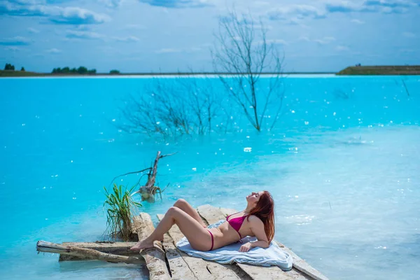 Una bella donna dai capelli rossi in un seducente bikini cremisi si siede di fronte a un lago turchese insolito. Un miracolo della natura, acqua limpida blu brillante. Cenere discarica, stagno avvelenato . — Foto Stock