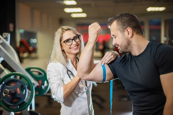 Badanie lekarskie. Atrakcyjny lekarz mierzy objętość bicepsów sportowców. Kobieta lekarz monitoruje przyrost mięśni u mężczyzny w siłowni — Zdjęcie stockowe