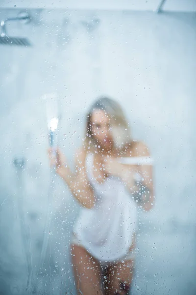 Uma menina delgada em uma camiseta branca molhada toma um banho, derrama água no copo. Janela com gotas de água antes da menina. Chuva cai no vidro da janela com o rosto da menina. Tratamento de spa de banho e higiene. — Fotografia de Stock