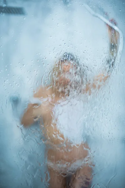 Ein schlankes Mädchen im weißen nassen T-Shirt duscht, gießt Wasser auf das Glas. Fenster mit Wasser fällt vor Mädchen. Regentropfen auf Fensterglas mit Mädchengesicht. Dusche und Hygiene-Wellness-Behandlung. — Stockfoto