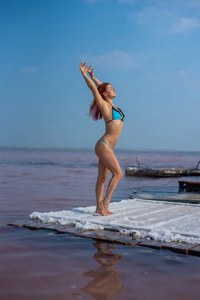 Piękna kobieta w uwodzicielskim niebieskim bikini stoi na drewnianej tratwie pokrytej solą. Słone jezioro jest różowe. Wpłata i ekstrakcja soli jadalnej. Cud natury.. — Zdjęcie stockowe