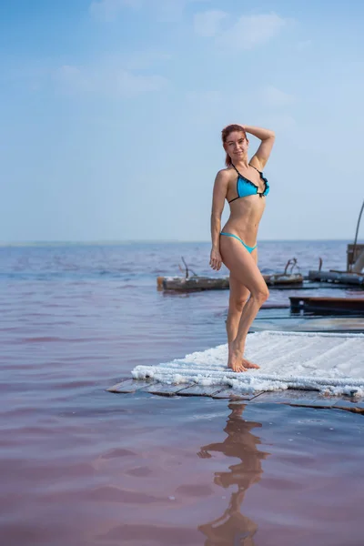 Una hermosa mujer en un seductor bikini azul se encuentra en una balsa de madera cubierta de sal. El lago de sal es rosado. Depósito y extracción de sal comestible. Milagro de naturaleza .. —  Fotos de Stock