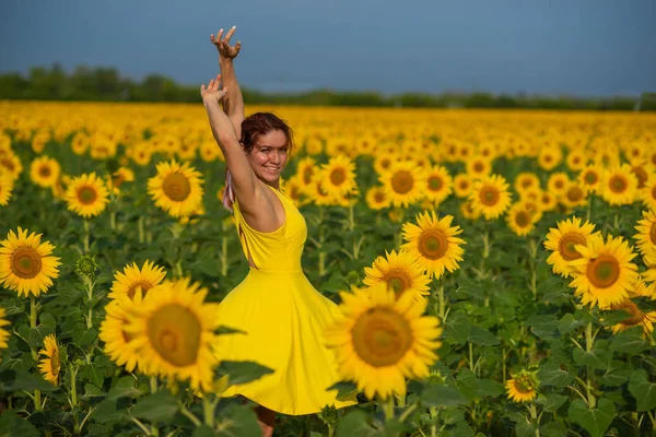 Rothaarige Frau in einem gelben Kleid, die mit erhobenen Händen in einem Sonnenblumenfeld tanzt. schöne Mädchen im Rock Sonne genießt einen wolkenlosen Tag auf dem Land. rosa Haarsträhnen. — Stockfoto