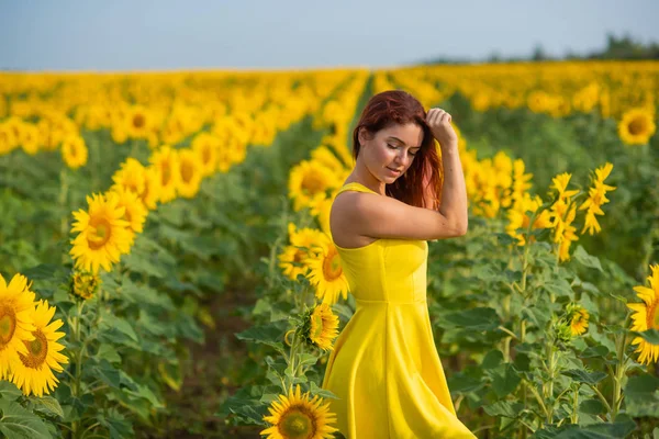一个穿着黄色衣服的红头发的女人站在向日葵的田野里。穿着裙子的漂亮女孩在乡下享受无云的一天。头发的粉红色锁. — 图库照片