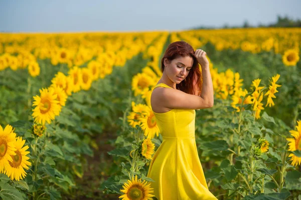 Eine rothaarige Frau in einem gelben Kleid steht auf einem Sonnenblumenfeld. schöne Mädchen im Rock Sonne genießt einen wolkenlosen Tag auf dem Land. rosa Haarsträhnen. — Stockfoto