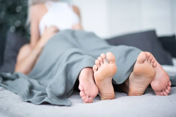 Close-up de pernas masculinas e femininas. O casal amoroso mente e abraça na cama sob um cobertor cinza. Sala de luz . — Fotografia de Stock