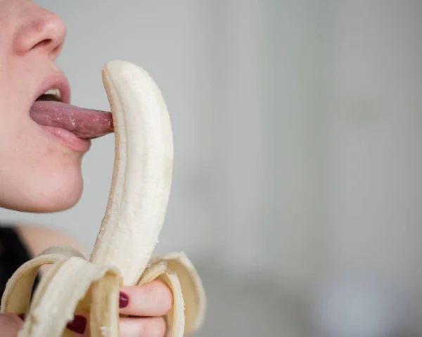 Close-up do rosto, lábios, língua de uma jovem, menina europeia lambendo uma grande banana amarela, doce . — Fotografia de Stock