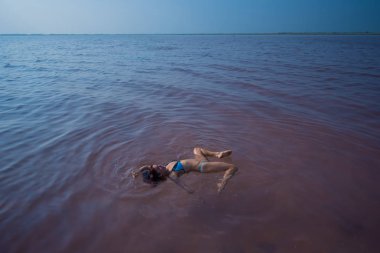 Mavi bikinili bir kadın tuz gölünde sırt larında yüzer. Doğanın mucizesi pembe göl. Tuz depozite ve çıkarma. Yoğun su yüzeye doğru itiyor. Maden suyunda tedavi prosedürleri.