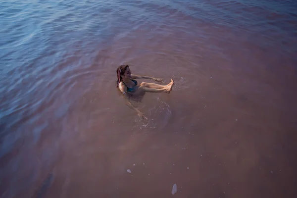 ある女性が塩湖で背中を泳いで笑う。自然ピンクの湖の奇跡。塩の堆積と抽出。表面に押し出す密な水。ミネラルウォーターの治療手順 — ストック写真