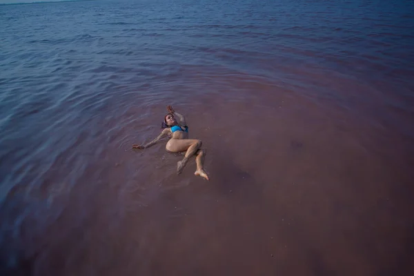 Žena v modrých bikinách jí na zádech plaví v solném jezeře. Zázrak přírody růžového jezera. Uložení a extrakce soli. Hustá voda tlačí na hladinu. Terapeutické postupy v minerální vodě. — Stock fotografie