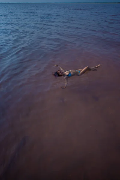 Una donna in bikini blu nuota sulla schiena in un lago salato. Miracolo della natura lago rosa. Deposito ed estrazione del sale. Acqua densa che spinge in superficie. Procedure terapeutiche nelle acque minerali . — Foto Stock