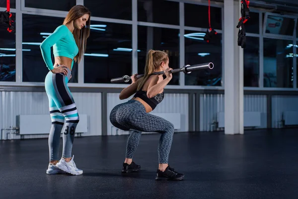 Young athletic girl doing barbell squats in the gym under the supervision of a trainer. A female crossfit coach teaches a ward to squat with a bar. Personal training. Exercises on the buttocks. — Stock Photo, Image